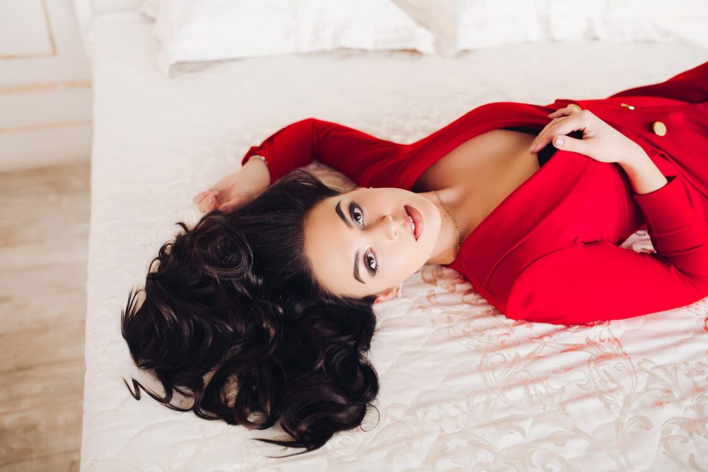Fată sexy în ținută roșie întinsă pe pat, cu ochi căprui.